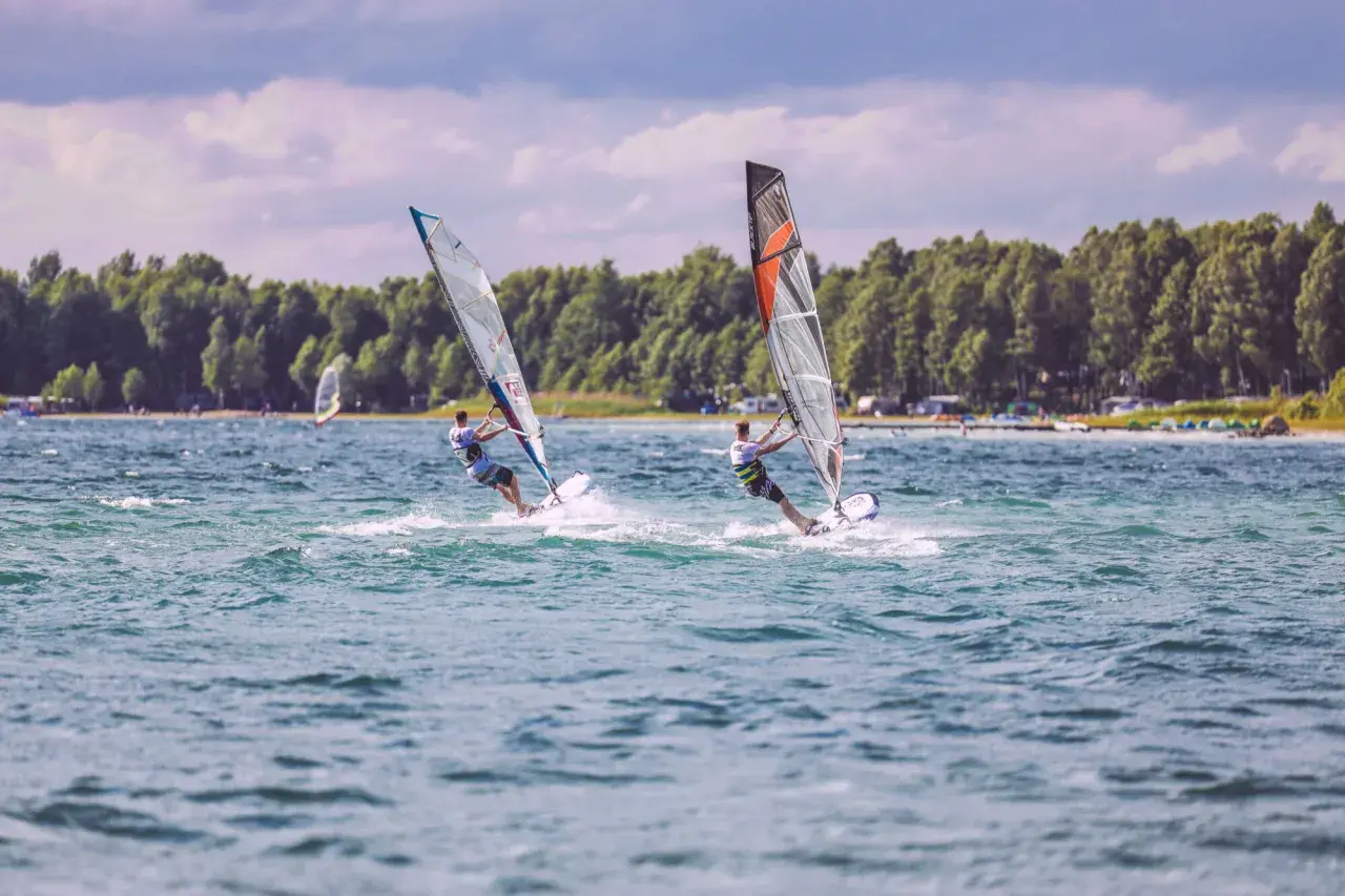 Dwóch surferów na desce z żaglem ściga się po falach. Idealne miejsce na windsurfing Mazury, gdzie wiatr i woda tworzą niezapomniane wrażenia.