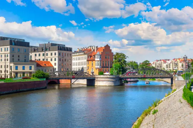 Widok na Odrę, rzekę płynącą przez Wrocław, z mostem i kolorowymi kamienicami w tle.